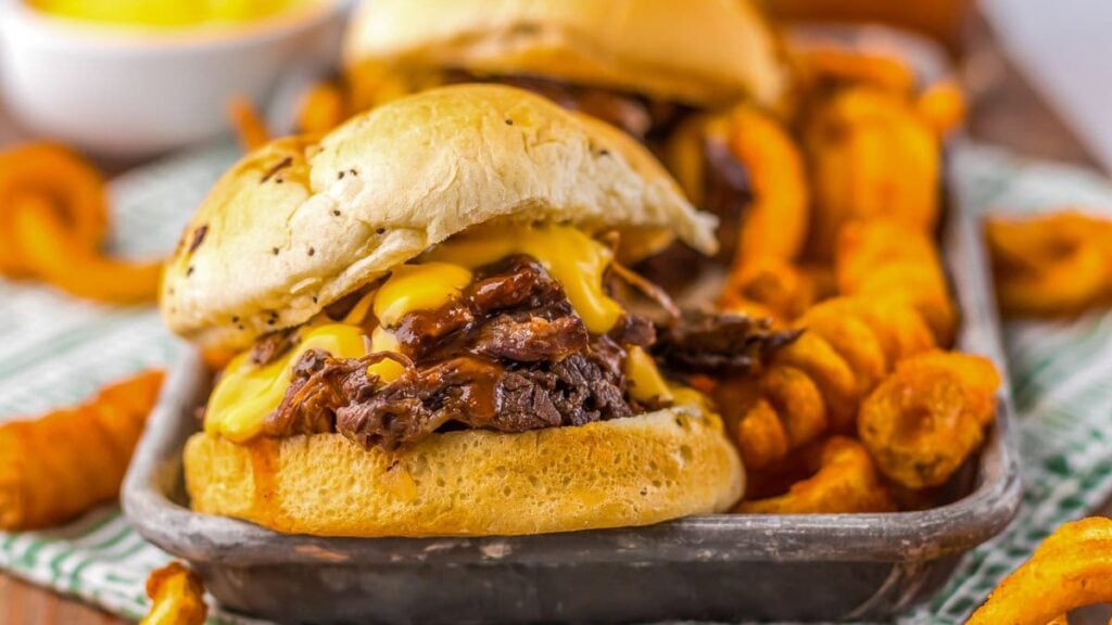 A shredded beef sandwich with melted cheese on a bun, served with curly fries on a tray.