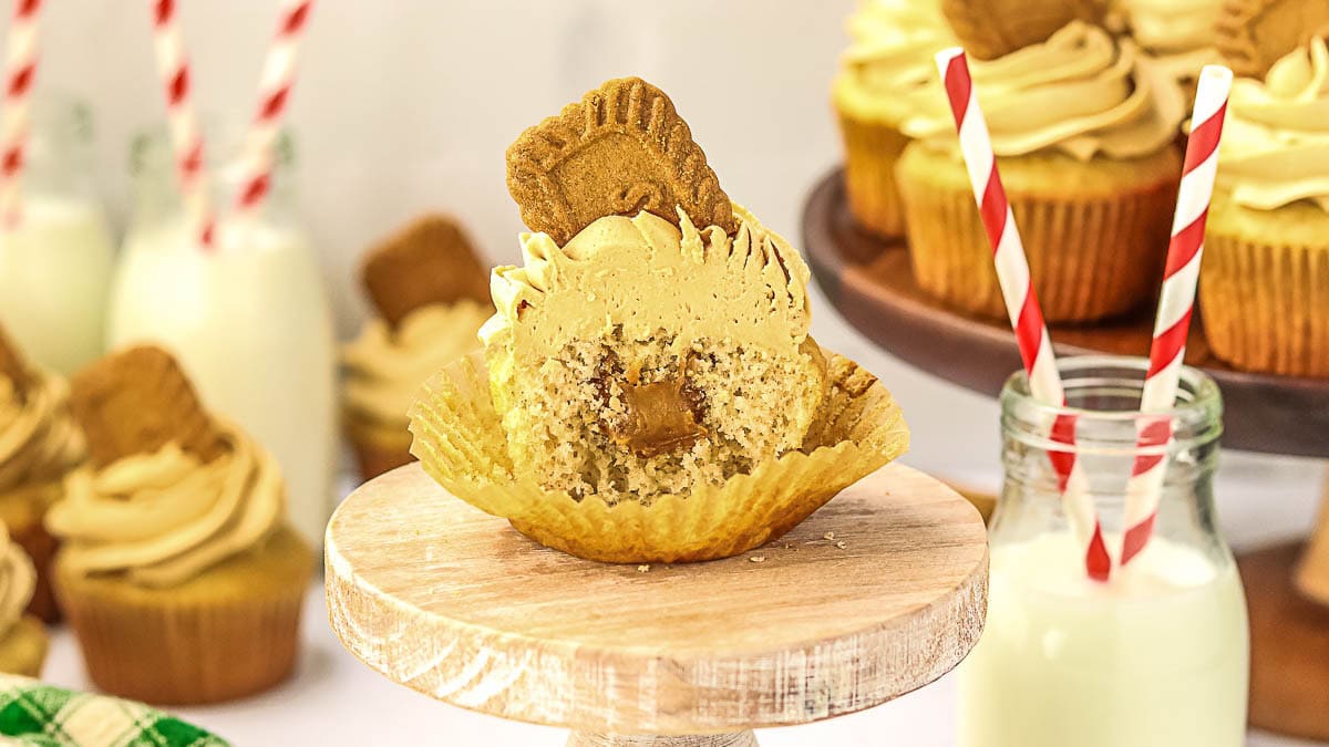 A cupcake with tan frosting and a cookie on top sits on a stand, partially unwrapped with a bite taken out to reveal a filling; cupcakes and milk bottles are in the background.