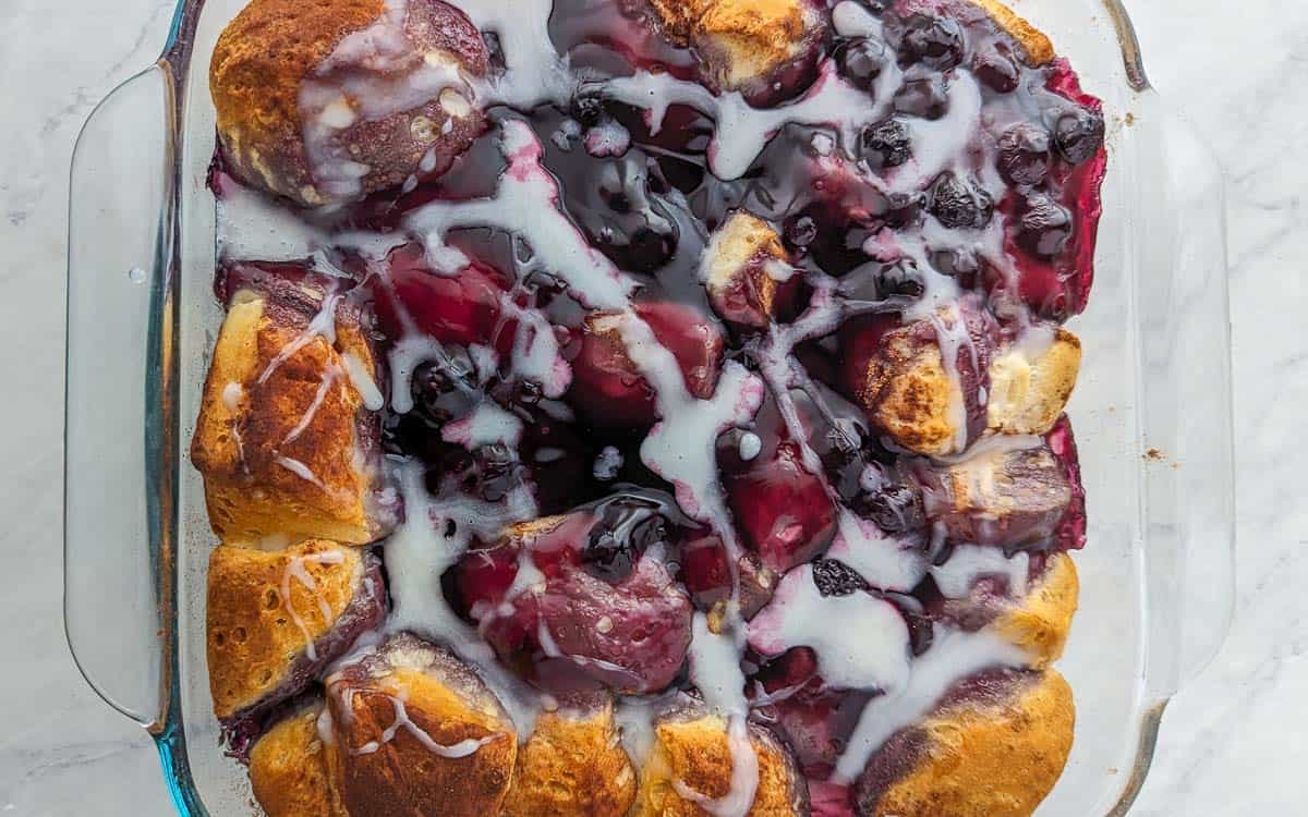 A square glass baking dish filled with a Blueberry Bubble Up topped with glazed dough pieces and drizzled with white icing.