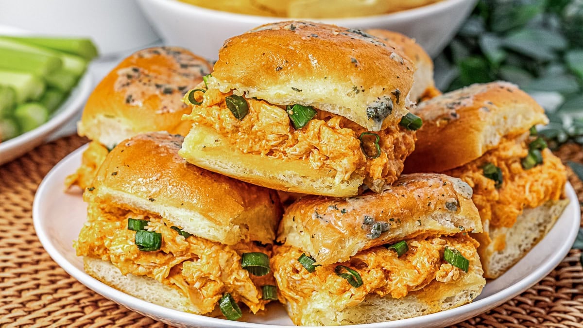 A plate of buffalo chicken sliders on seasoned buns, garnished with chopped green onions, is arranged on a white plate.