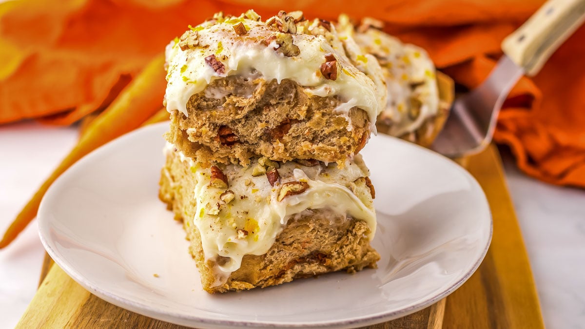 Two carrot cake bars with cream cheese frosting and chopped nuts on a white plate, set on a wooden board with an orange napkin and a serving spatula in the background.