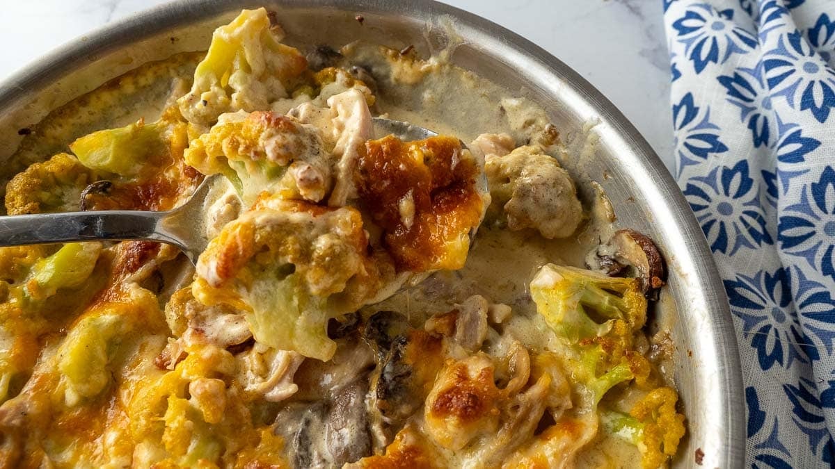A close-up of a cheesy baked casserole with cauliflower, mushrooms, and a creamy sauce in a metal dish, with a serving spoon lifting a portion.