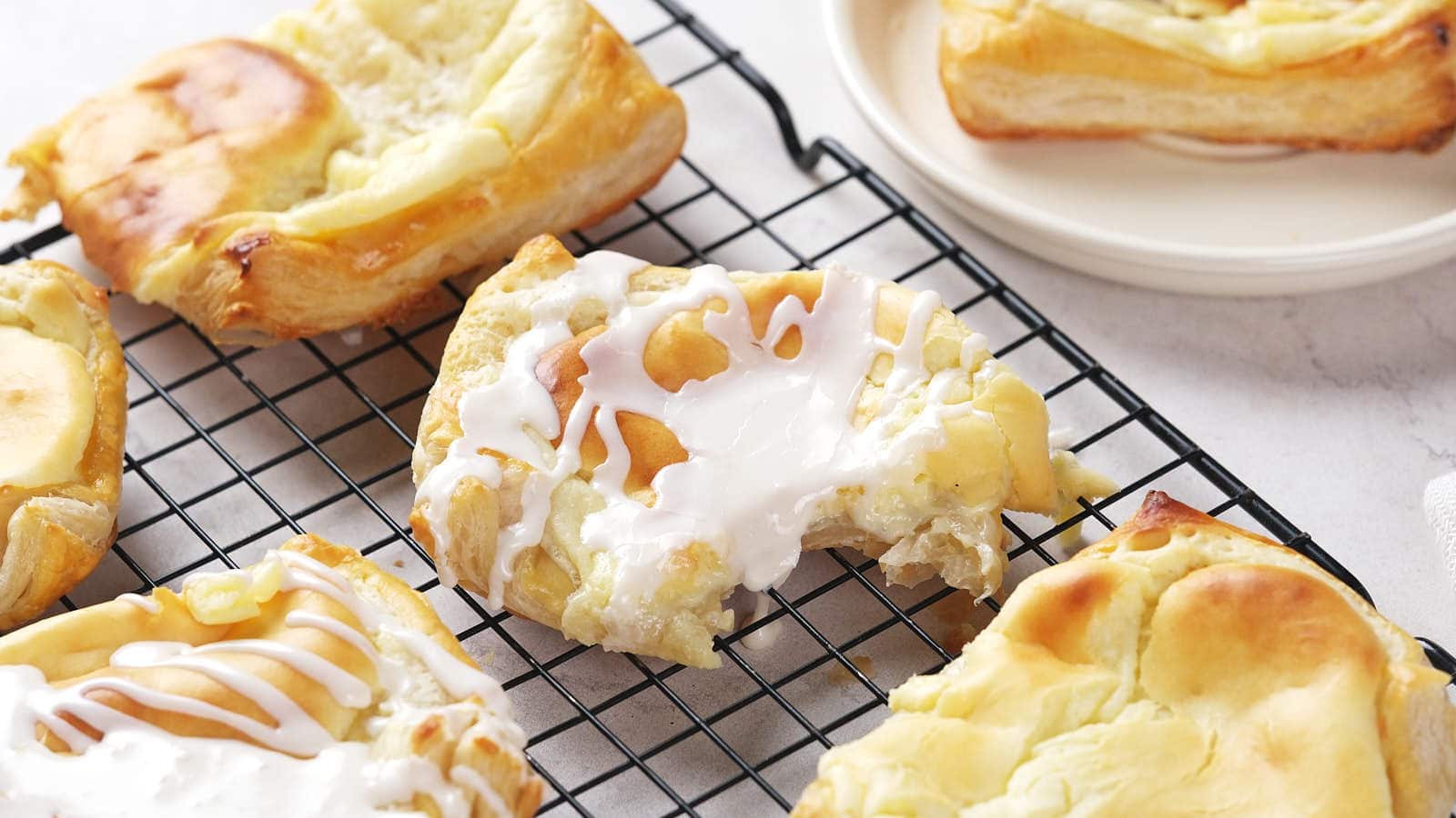 Cheese danish with icing are cooling on a black wire rack, with one pastry missing a bite.