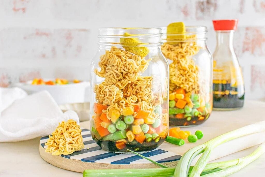 Two glass jars prepped for meal prep, filled with layers of uncooked ramen noodles, frozen mixed vegetables, and sauce, are placed on a striped board with green onions and a soy sauce bottle nearby.