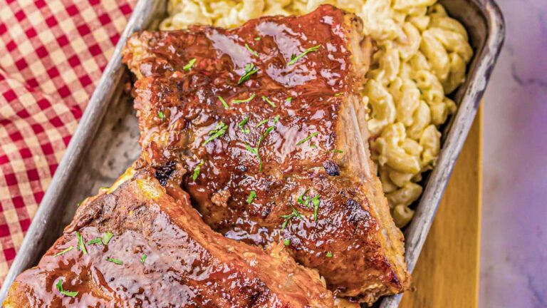 Two glazed barbecue ribs placed on top of creamy macaroni and cheese in a metal tray, with a red and white checkered cloth partially visible.