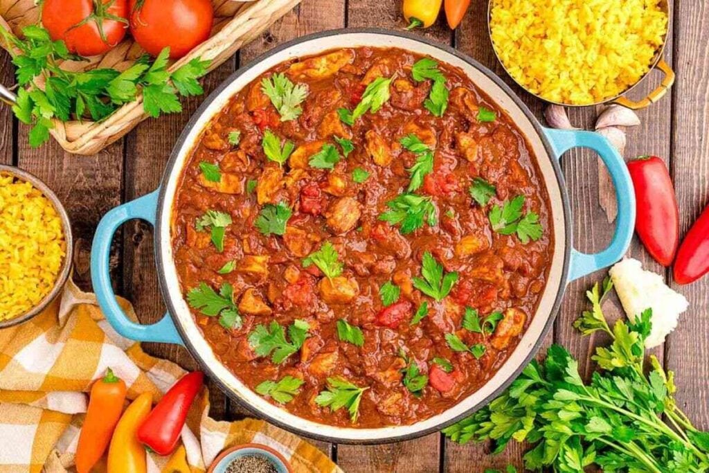 A pot of tomato-based stew with chunks of meat and vegetables, garnished with parsley, sits on a wooden table beside bowls of yellow rice, fresh herbs, and peppers.