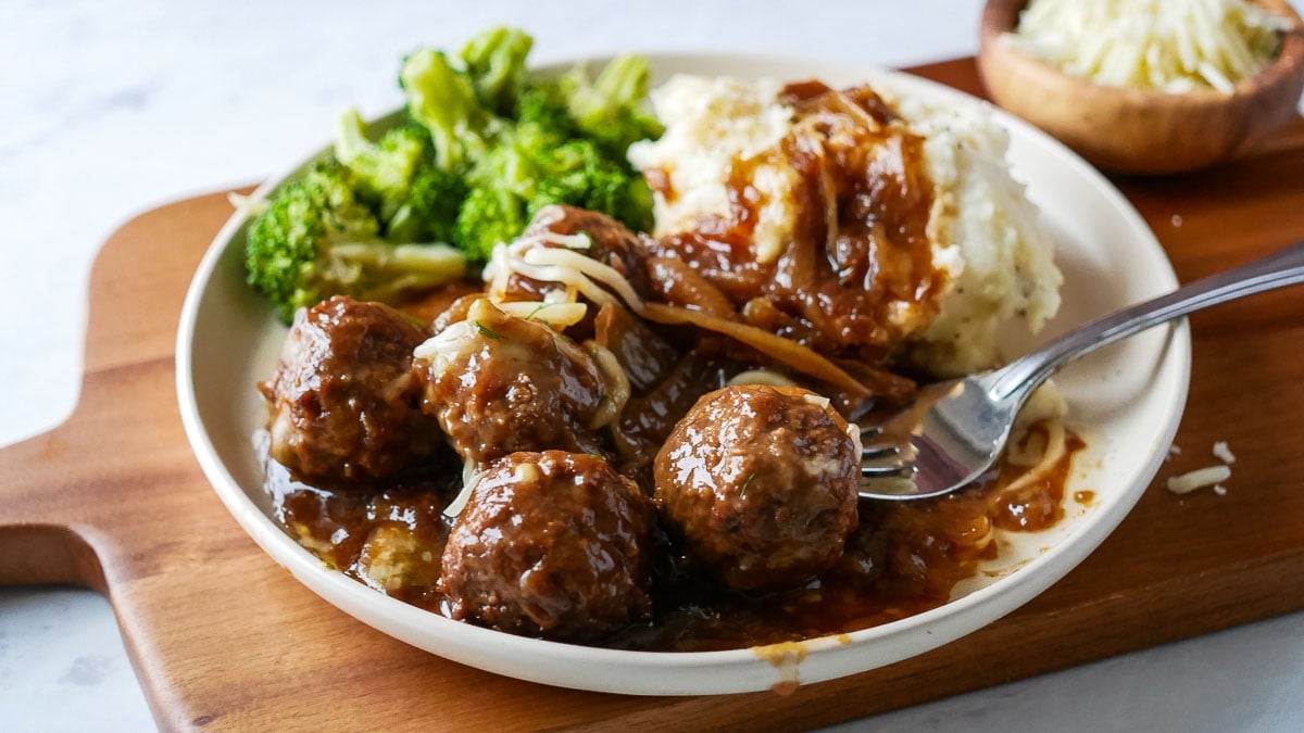 A plate of meatballs in gravy, mashed potatoes, and broccoli on a wooden board.