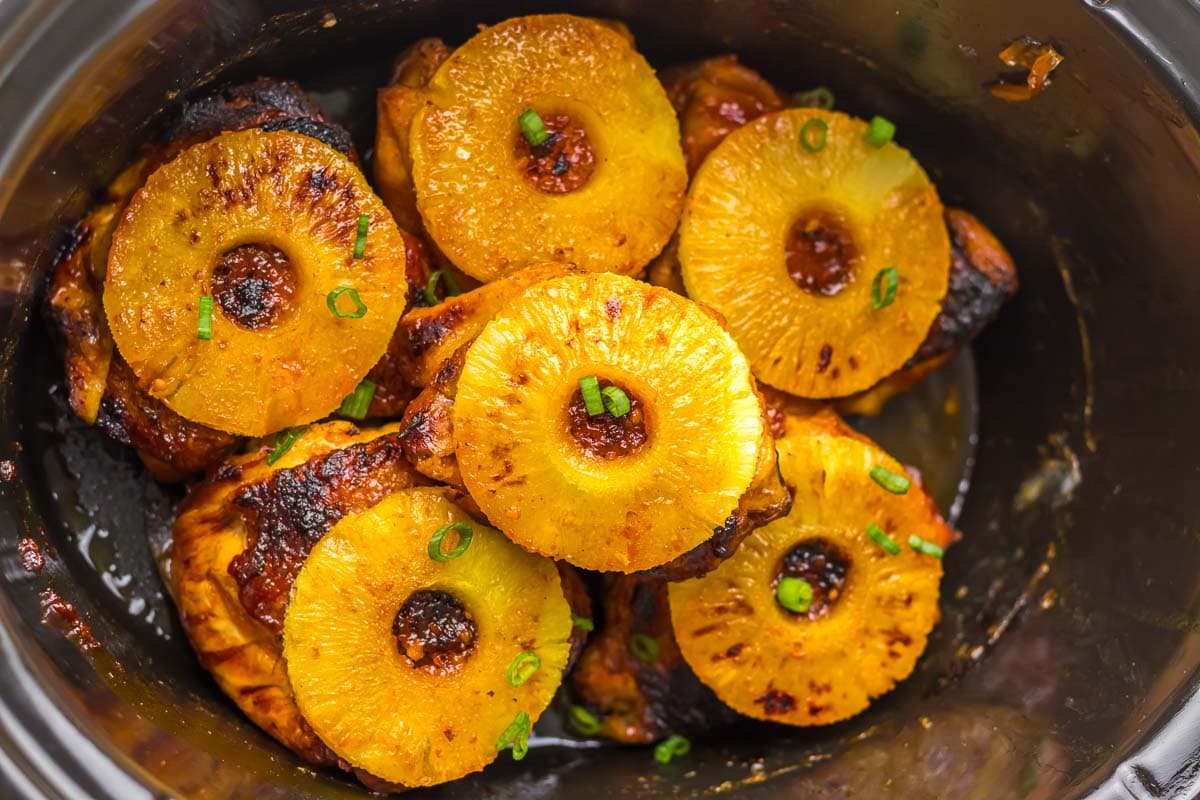 Pineapple slices arranged over cooked chicken pieces in a slow cooker.
