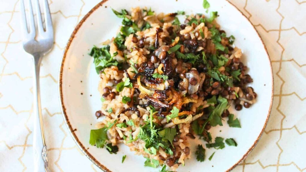 A white dish filled with cooked lentils and rice, topped with chopped cilantro and caramelized onions. A wooden spoon rests on a patterned napkin next to the dish.