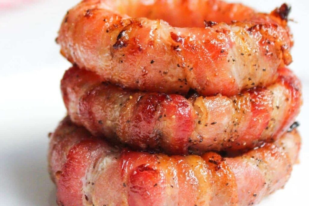 Three bacon-wrapped onion rings stacked on a white surface, showing crispy, cooked bacon on the outside.
