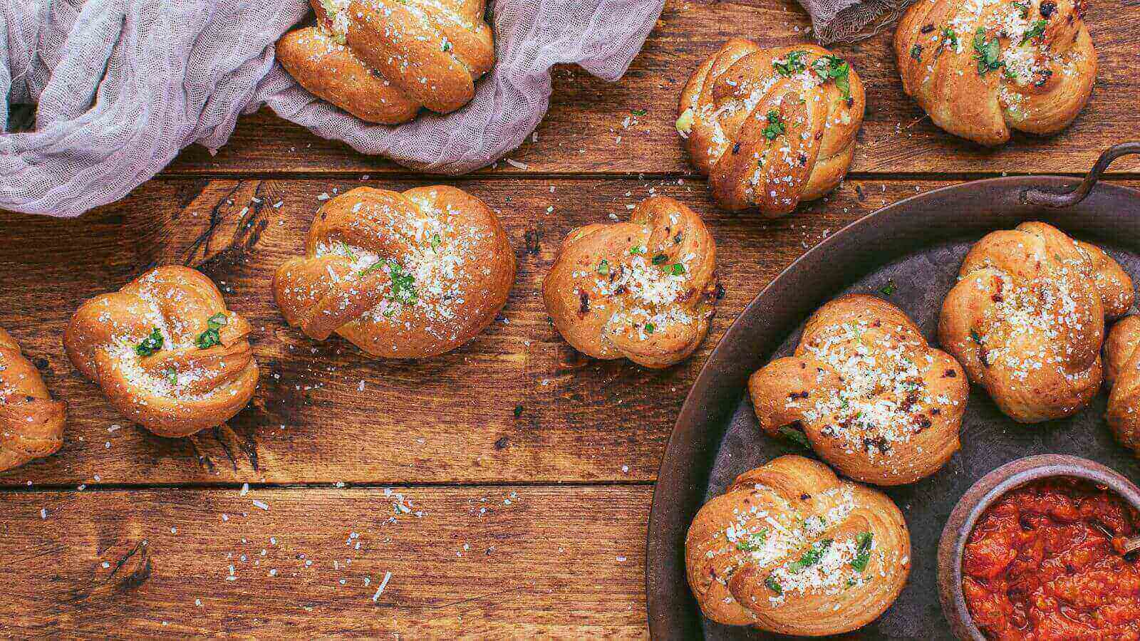Garlic knots sprinkled with herbs and grated cheese are displayed on a wooden surface, alongside a bowl of tomato sauce.