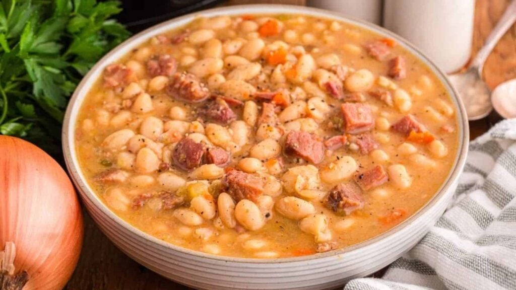 A bowl of hearty white bean soup with chunks of ham, surrounded by a fresh onion, parsley, and kitchen utensils.