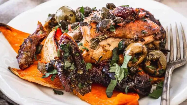 A plate of roasted chicken with olives, capers, dried fruit, and herbs served on top of roasted sweet potato wedges, with a fork on the side.