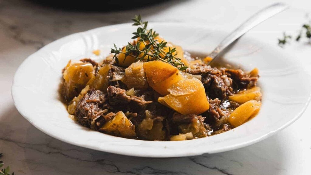 A white bowl filled with beef stew containing chunks of meat, potatoes, and herbs, with a sprig of thyme on top and a spoon in the bowl.