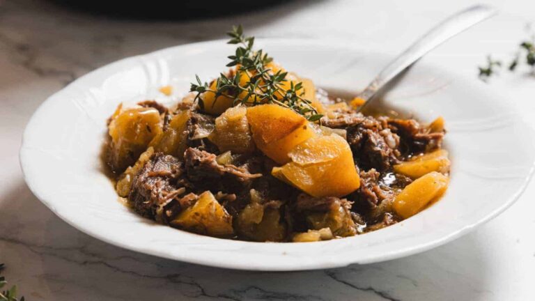 A white bowl filled with beef stew containing chunks of meat, potatoes, and herbs, with a sprig of thyme on top and a spoon in the bowl.