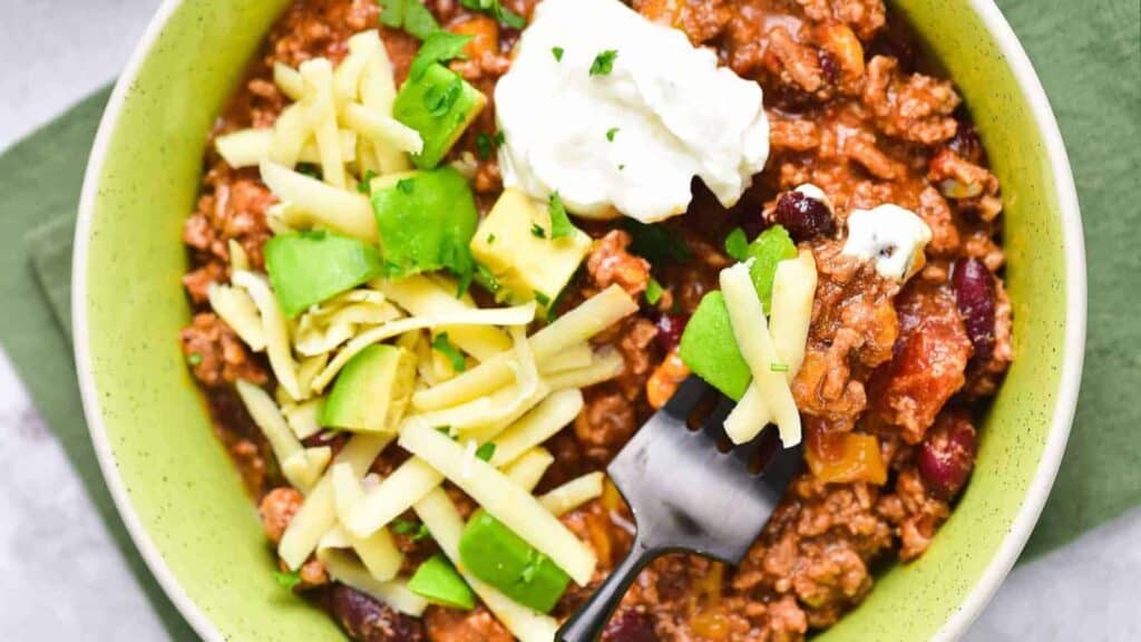 A bowl of chili topped with shredded cheese, diced avocado, and a dollop of sour cream, with a fork resting in the bowl. The food is served in a light green bowl on a green napkin.