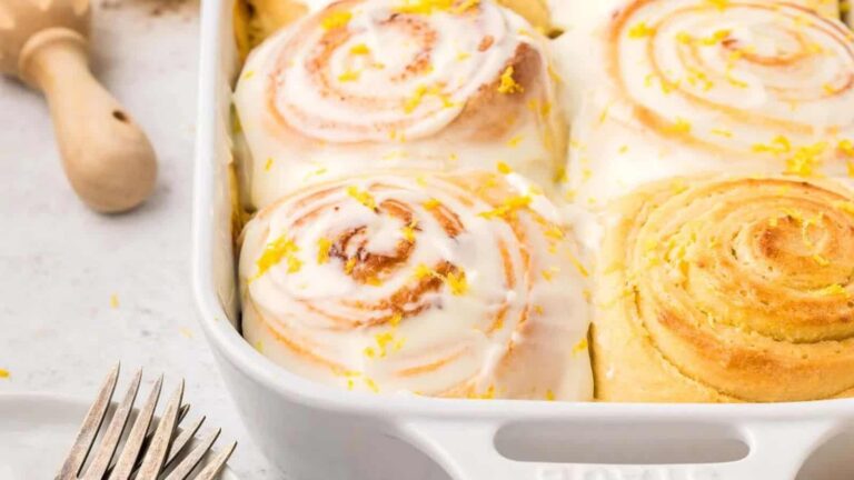A white baking dish with iced cinnamon rolls topped with lemon zest, next to a fork and a wooden utensil on a light countertop.