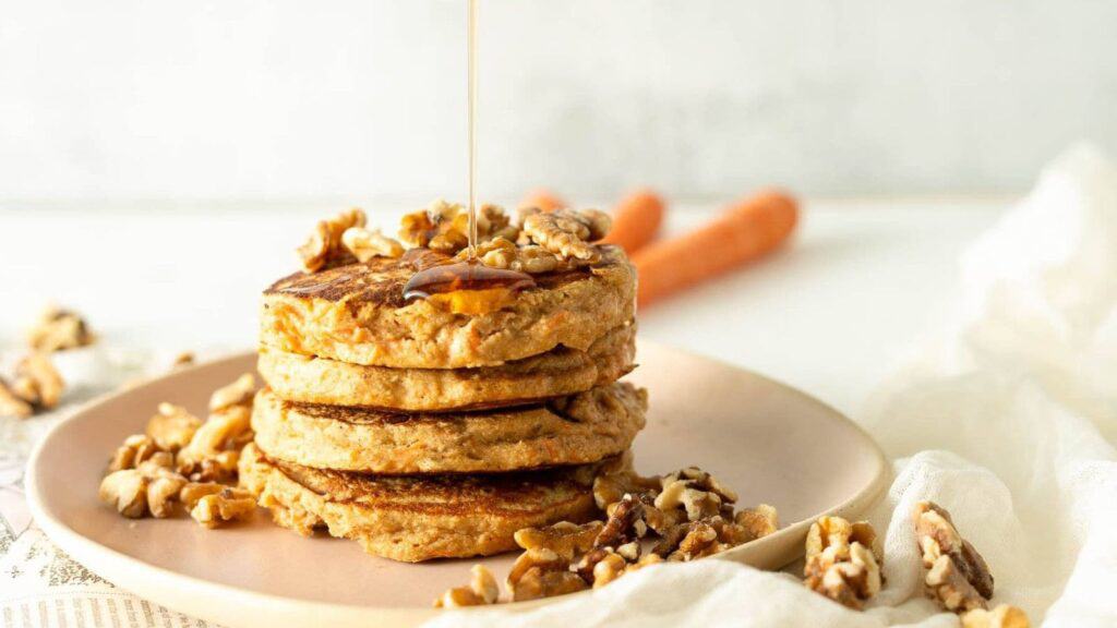 A stack of pancakes topped with chopped walnuts is being drizzled with syrup on a light-colored plate.