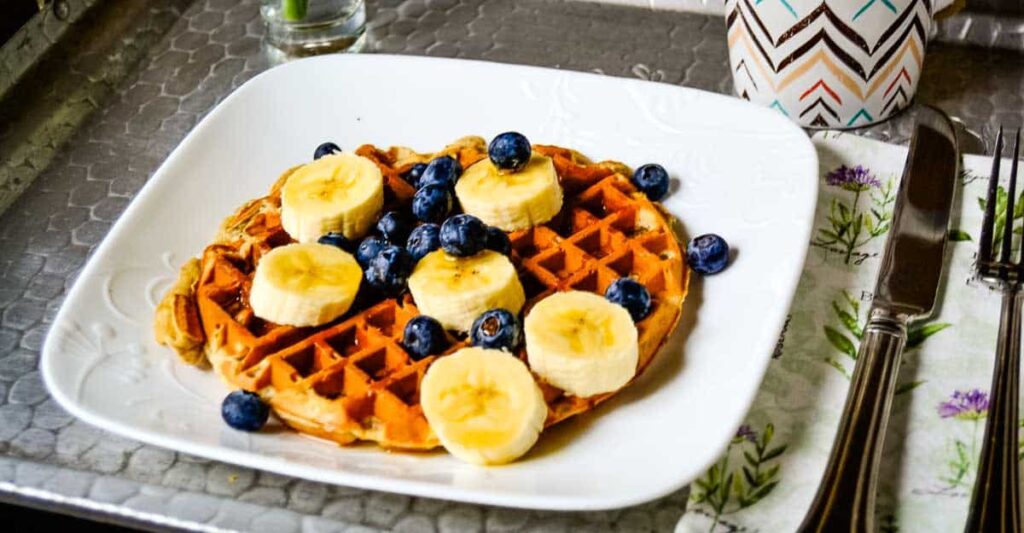 A waffle topped with banana slices and blueberries is served on a white plate with a fork and knife on the side.