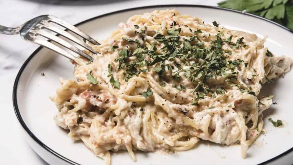 A plate of creamy spaghetti with shredded chicken, topped with chopped parsley and black pepper.