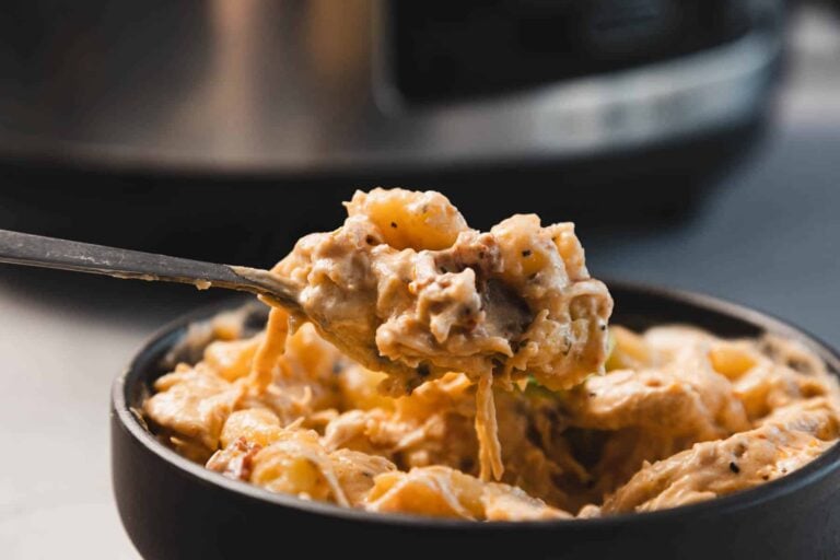 A spoon lifts a serving of creamy pasta with cheese and visible pieces of meat from a black bowl, with a kitchen appliance in the blurred background.