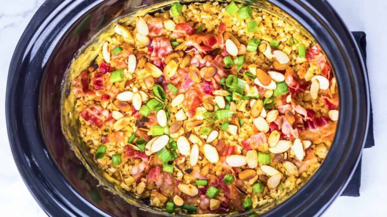 Crockpot filled with a cooked dish topped with sliced almonds, chopped green onions, and pieces of bacon.