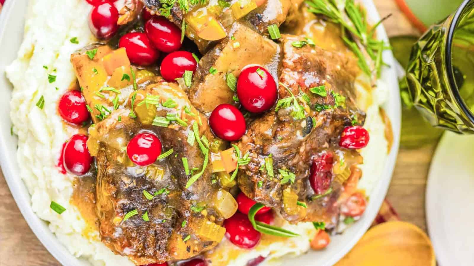 Braised meat with gravy, fresh cranberries, and herbs served over mashed potatoes on a white plate.