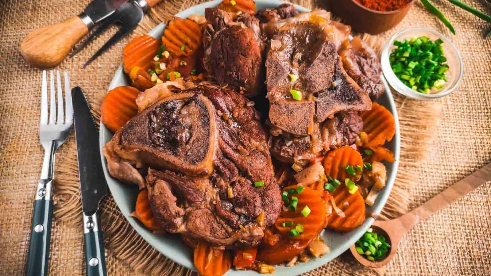 A plate of cooked lamb chops sits on a bed of sliced, cooked carrots. The dish is garnished with chopped green onions. Utensils, a wooden spoon with spices, and a small bowl of more green onions are nearby on a burlap cloth surface.