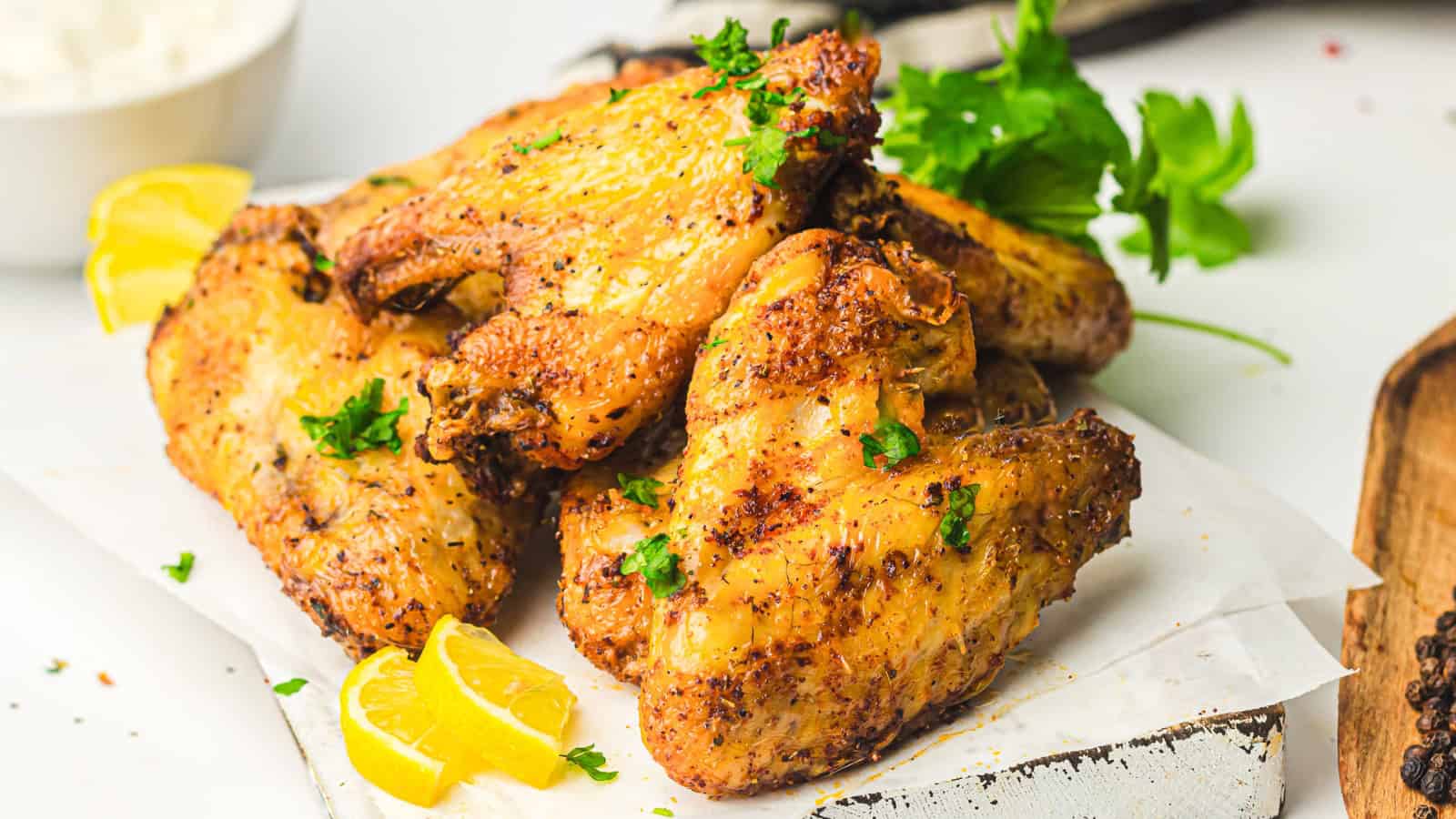 Close-up of a plate of seasoned and grilled chicken wings garnished with herbs. Lemon wedges are visible in the background, adding a hint of color. The wings appear crispy and well-cooked, resting on a dark plate.