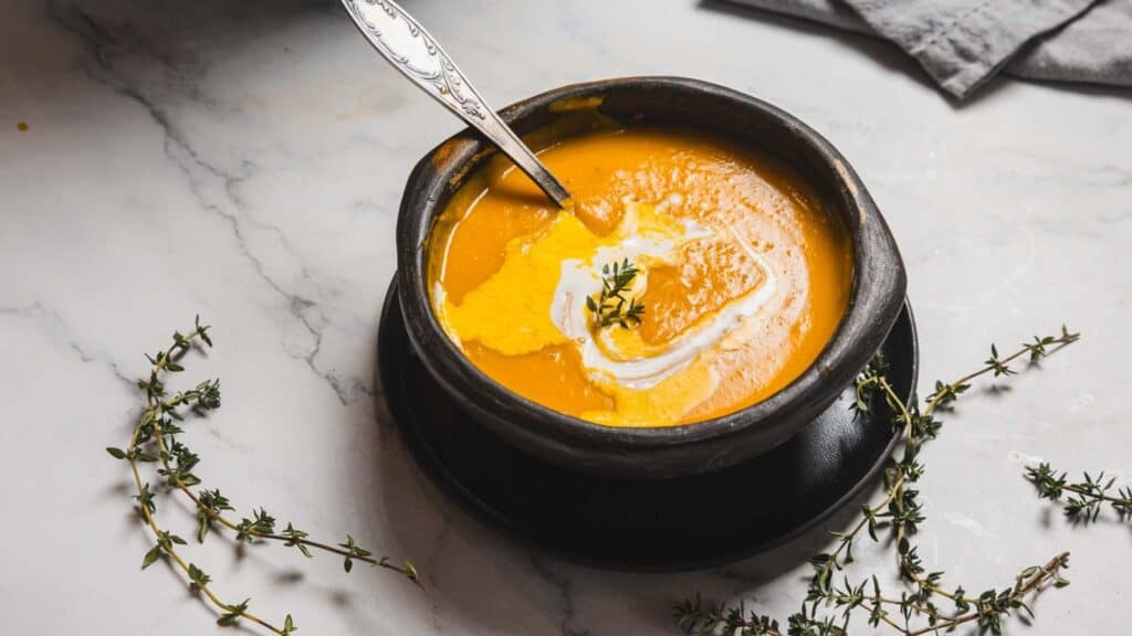 A bowl of creamy orange soup is placed on a black plate, garnished with a swirl of cream and a sprig of herb. A spoon rests inside the bowl. Small sprigs of thyme are scattered on the marble surface nearby.