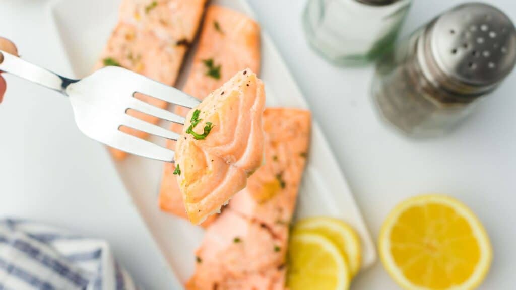 Four grilled salmon fillets garnished with fresh dill and lemon slices are arranged on a white oval platter. The fillets are seasoned with herbs and pepper. The edge of a striped cloth and a serving of broccoli are partially visible.