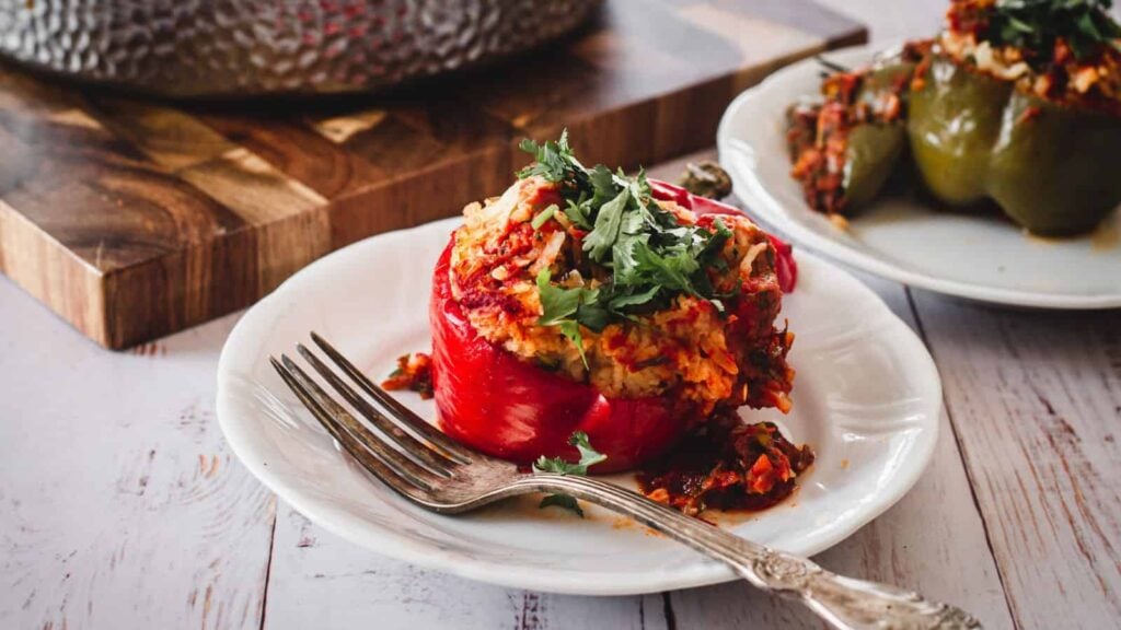 A stuffed red bell pepper filled with rice and vegetables, topped with herbs, is served on a white plate with a fork. Another stuffed pepper is visible in the background.