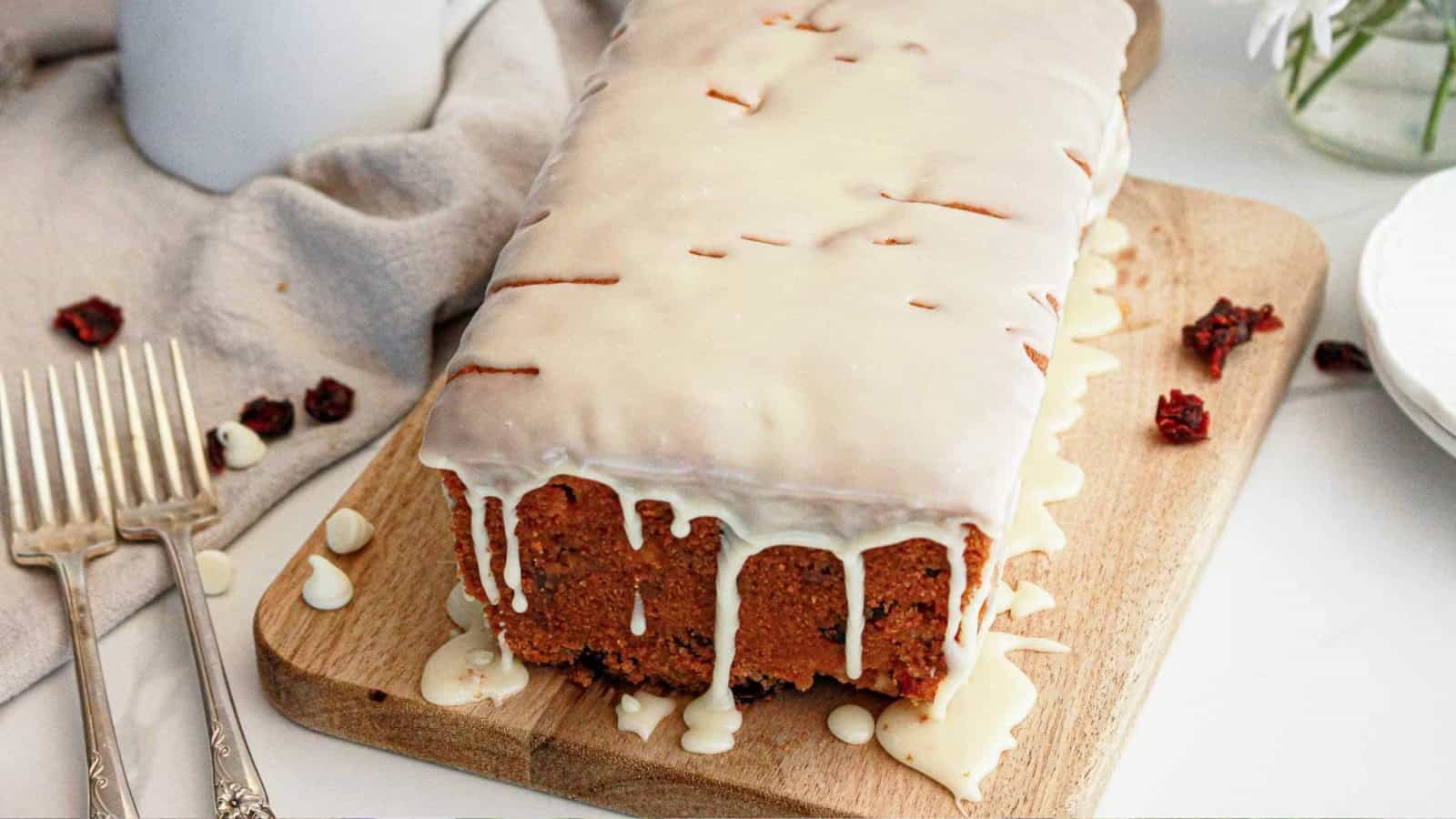 A loaf cake with white icing drips on a wooden board, next to two forks and some dried fruit pieces.