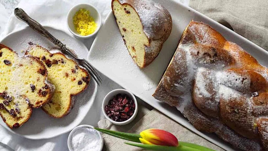 A braided loaf dusted with powdered sugar is partially sliced on a platter, with slices served on a plate beside bowls of dried cranberries, lemon zest, and powdered sugar.