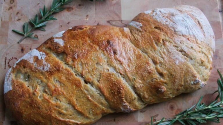 A loaf of rustic bread with a golden crust sits on a wooden surface, garnished with fresh rosemary sprigs.