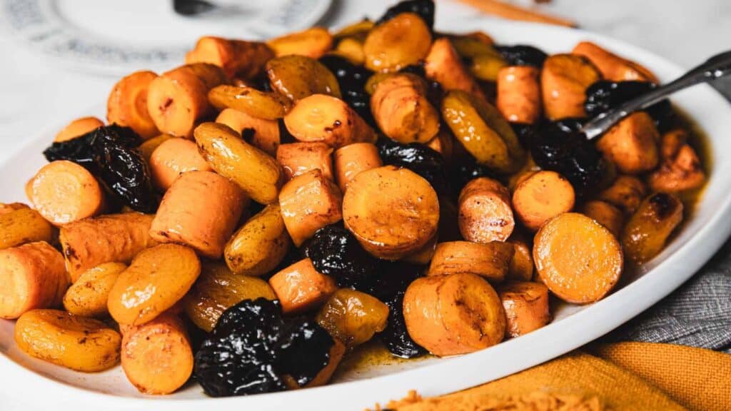 A white platter filled with roasted carrots, sweet potatoes, and prunes, served with a fork on the side.