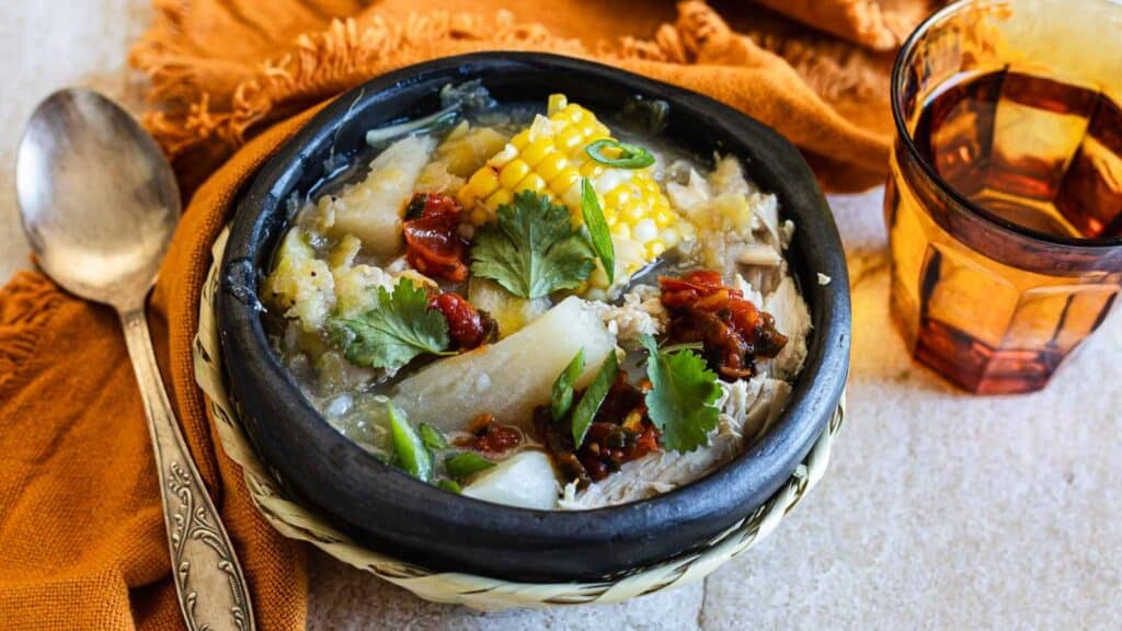 A bowl of soup containing corn on the cob, cilantro, and various vegetables is placed on a woven mat. A spoon lies to the left, and an amber-colored glass with a beverage is to the right on a light-colored surface. An orange cloth napkin is underneath—this isn't just any soup; it's inspired by Grandma's Chicken Recipes.