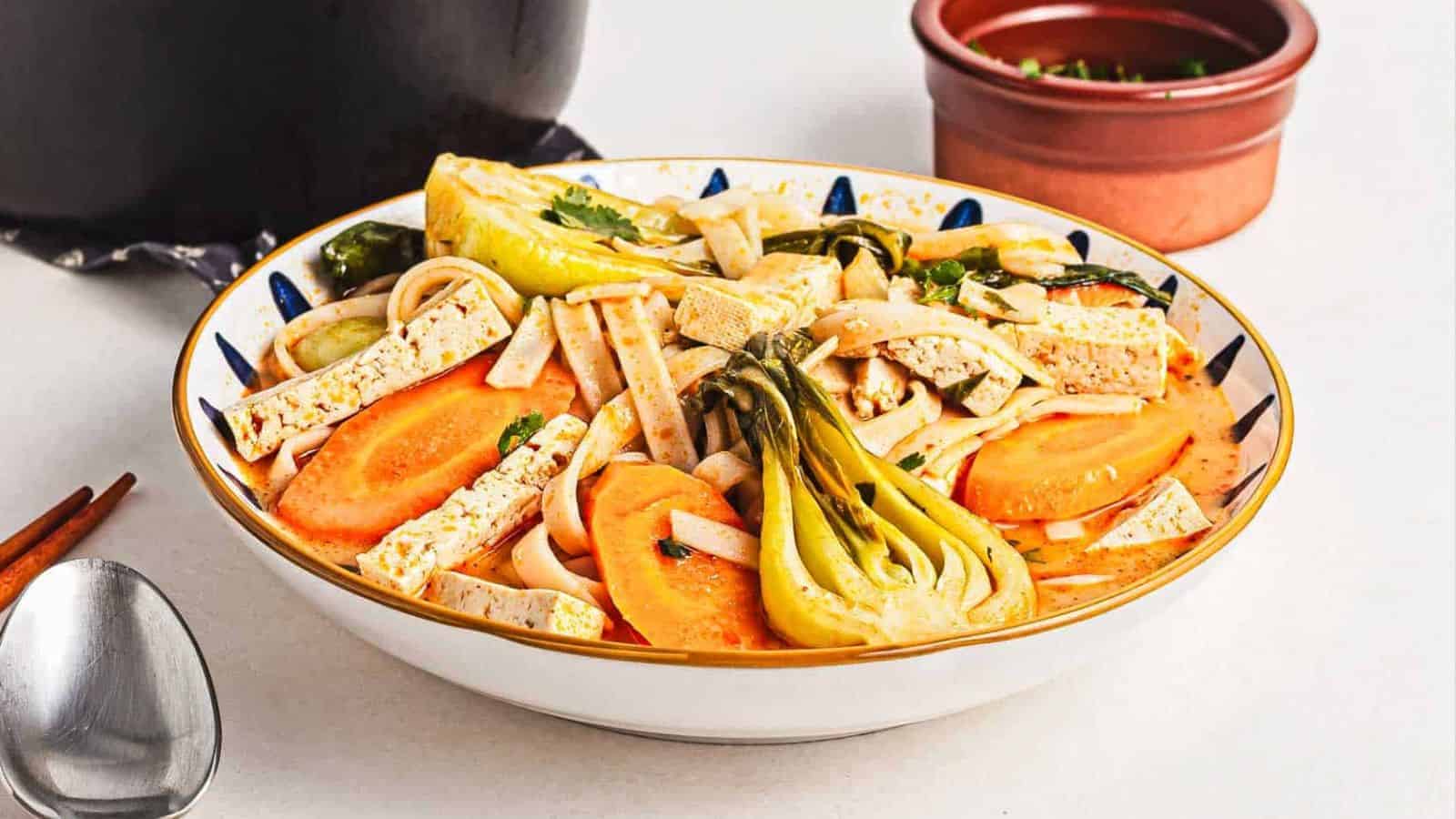 A bowl of noodle soup with tofu strips, baby bok choy, carrot slices, and fresh herbs, set on a white surface with a spoon nearby and a small bowl of chopped greens in the background.