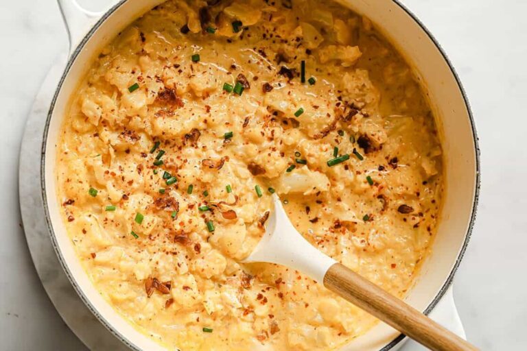 A pot of creamy cauliflower topped with chopped chives and red pepper flakes, with a wooden spoon resting inside.