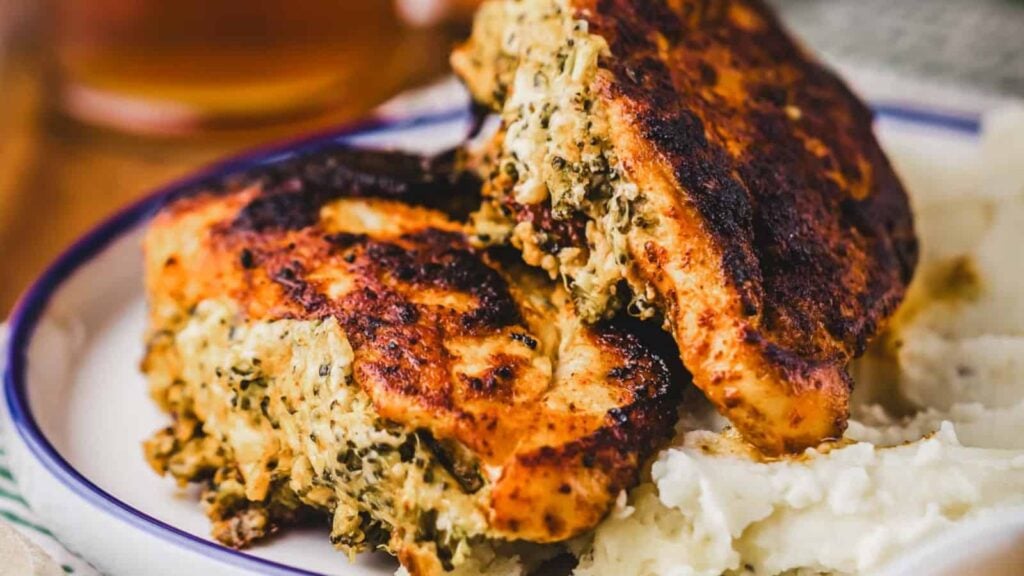 Two pieces of stuffed and grilled chicken breast with a browned, crispy exterior are served on a plate next to a portion of mashed potatoes. The chicken stuffing appears to contain herbs and cheese.