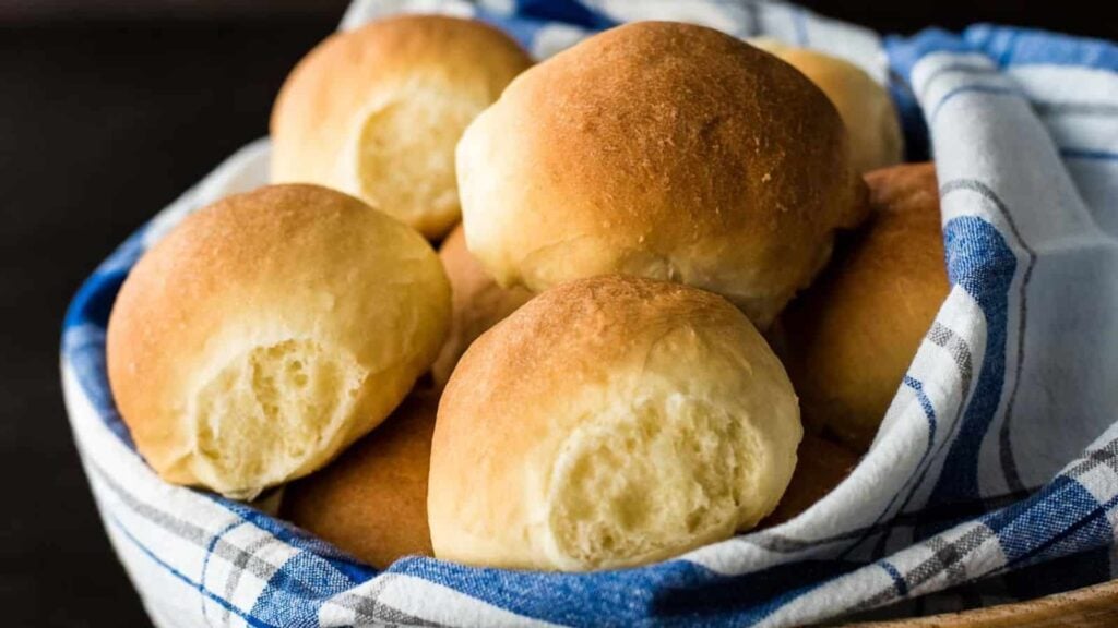 A basket lined with a blue and white cloth holds several round, golden-brown dinner rolls.