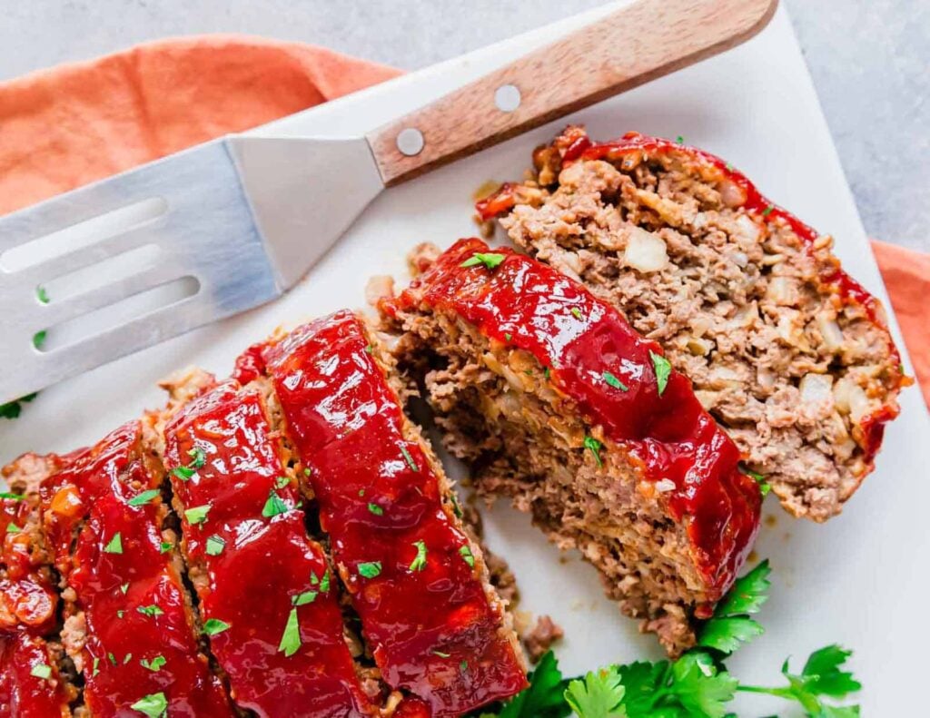 Sliced meatloaf with a tomato glaze on a white cutting board, garnished with parsley, next to a metal spatula and an orange napkin.