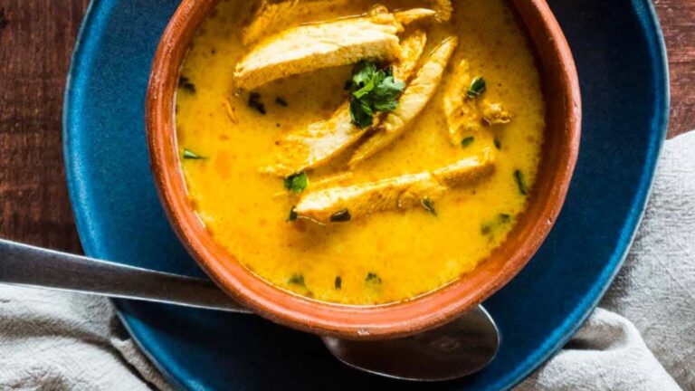 A bowl of yellow curry soup with sliced chicken and herbs, served on a blue plate with a spoon and napkin beside it.