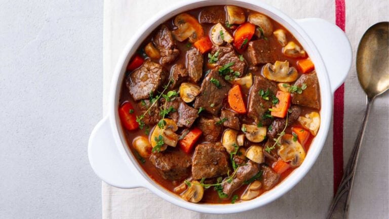 A white bowl filled with beef stew, featuring chunks of beef, sliced mushrooms, carrots, and herbs in a brown broth, placed on a beige cloth next to a spoon.
