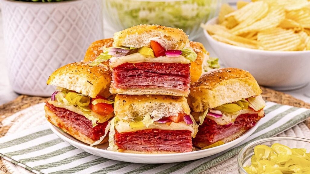 A plate of stacked Italian sub sandwiches filled with deli meats, cheese, lettuce, tomatoes, onions, and peppers, with a bowl of potato chips in the background.