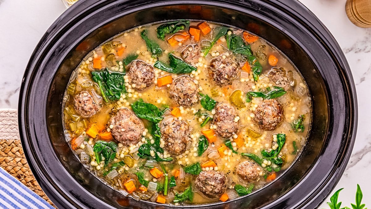 A slow cooker filled with soup containing meatballs, carrots, spinach, celery, pasta, and broth on a marble countertop.