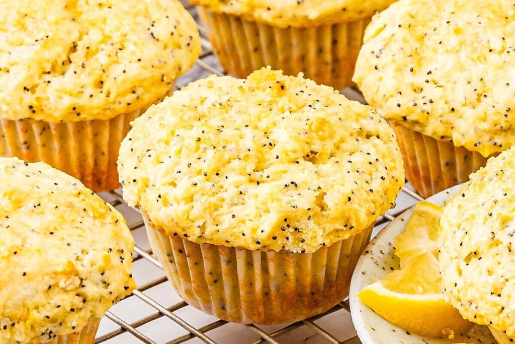 Cozy lemon poppy seed Easy Muffins rest on a cooling rack, with a sliced lemon on a small plate nearby.