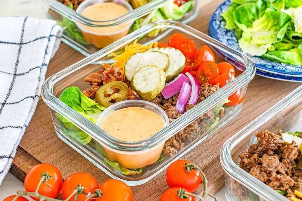 Perfect for meal prep, this glass container holds ground beef, lettuce, cherry tomatoes, pickles, red onions, jalapeños, shredded cheese, bacon bits, and a cup of creamy dressing.
