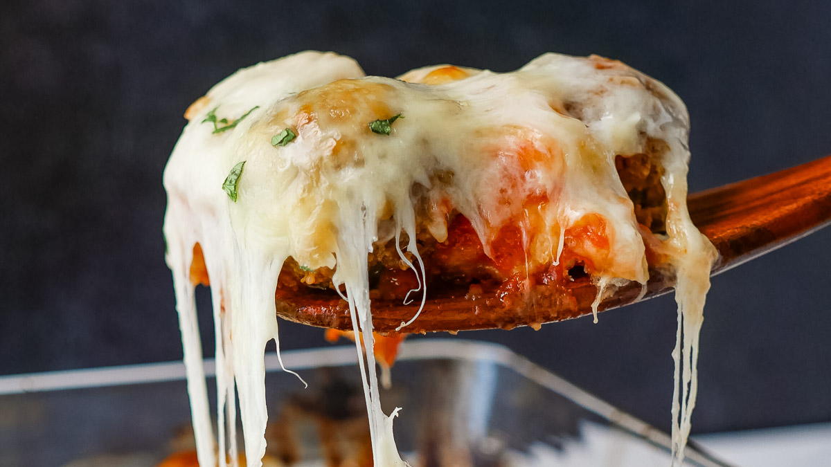 Close-up of a wooden spoon lifting a cheesy meatball from a glass baking dish, with melted cheese stretching and herbs on top.