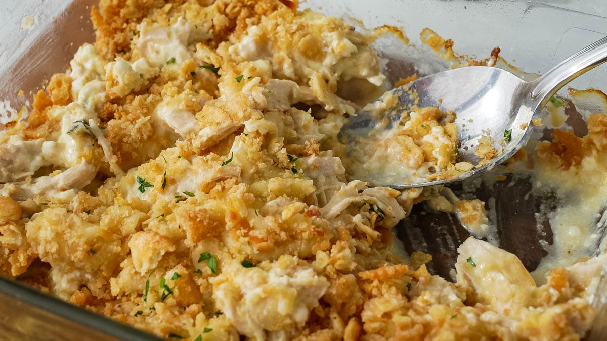 A dish of creamy baked chicken casserole and cheese topped with golden, crispy crackers, garnished with parsley, served on a glass dish with a fork.